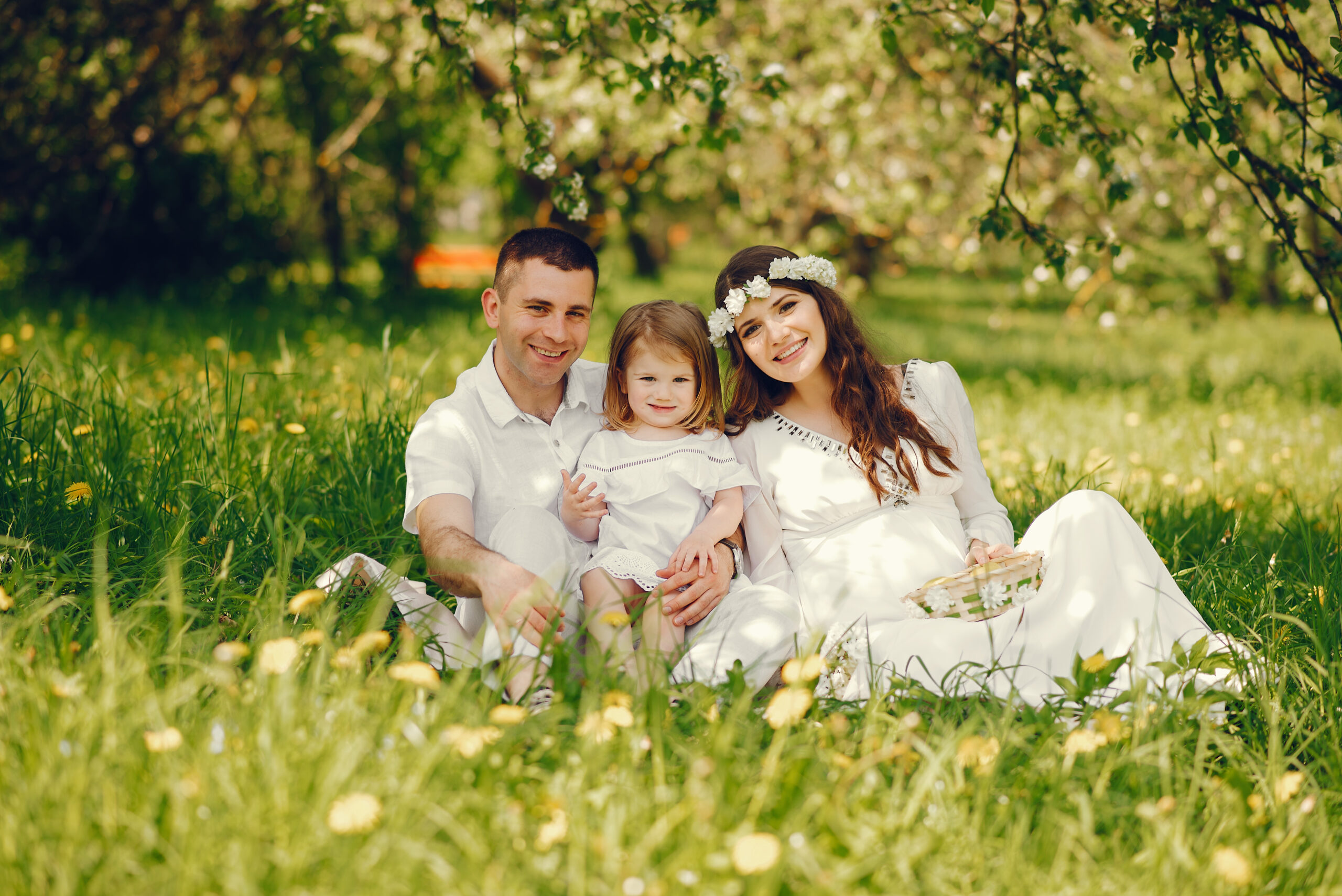 beautiful young pregnant girl in a long white dress and wreath on her head playing in a sunny summer park between the trees with her little daughter and husband