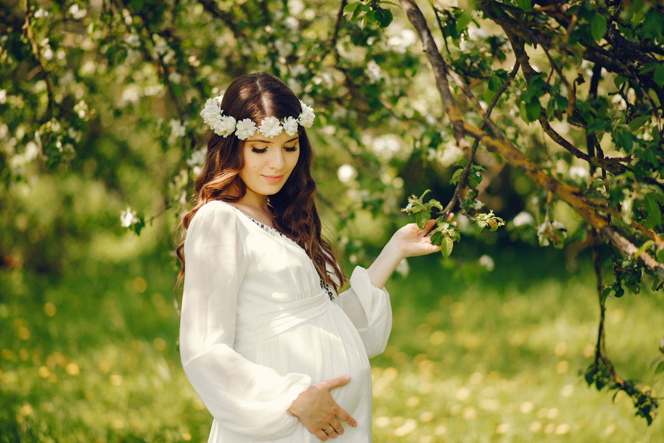 beautiful young pregnant girl in a long white dress and wreath on her head walking in a sunny summer park between the trees