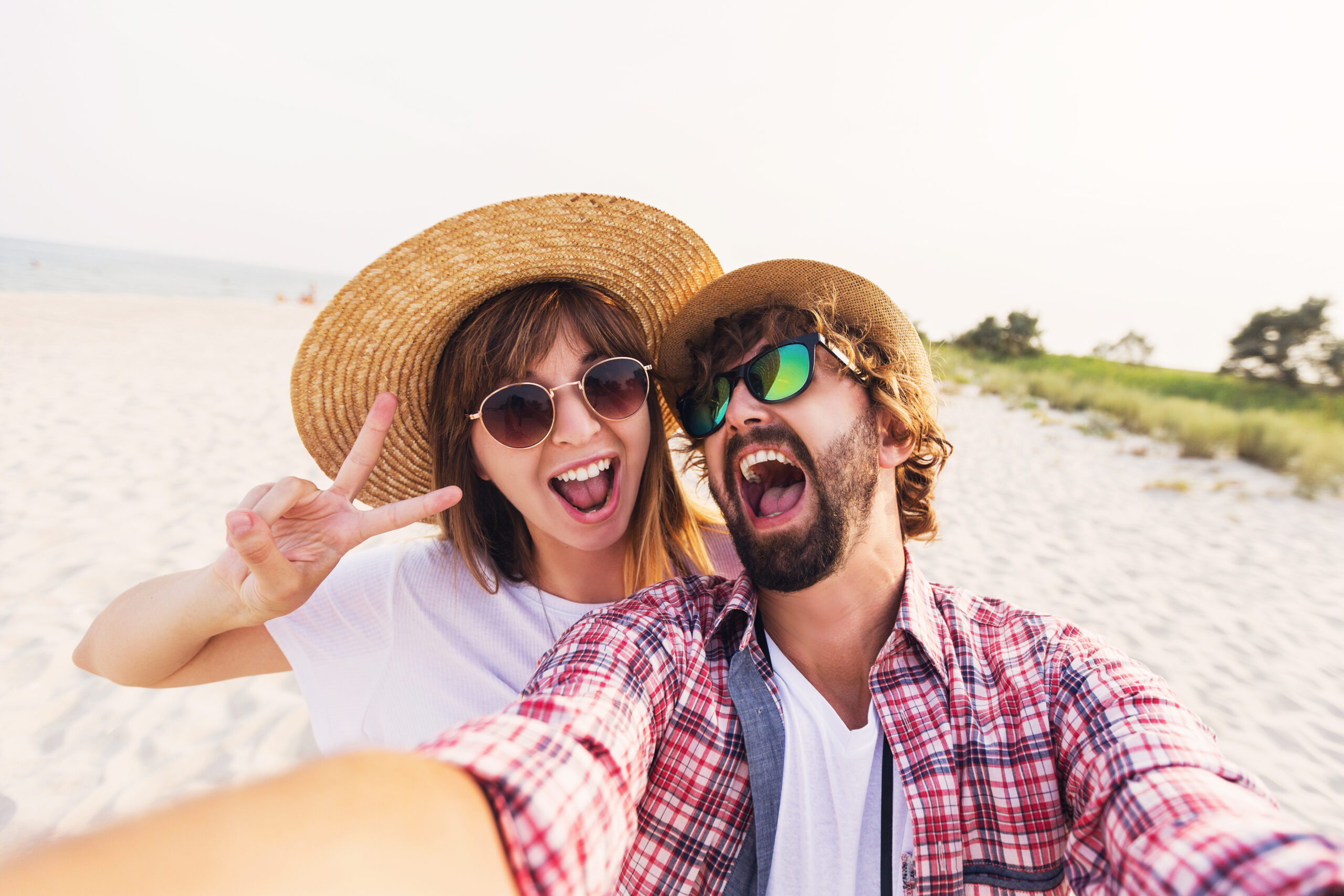 Happy   traveling couple in love taking a selfie on phone at the beach on a sunny summer day. Pretty girl and her handsome boyfriend with beard having fun, crazy emotional faces , laughing.