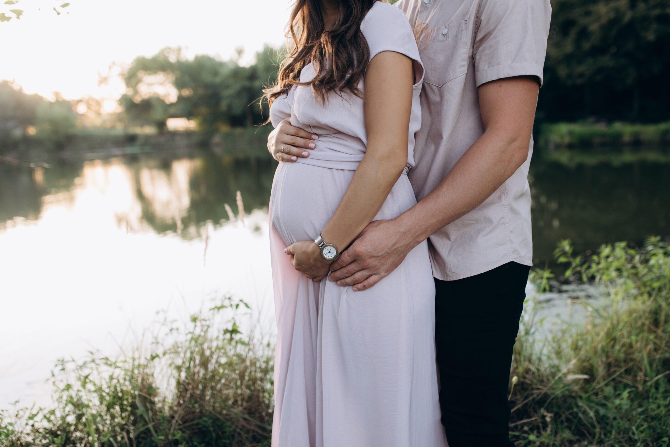 Man hugs beautiful pregnant woman tender standing before the lake in the rays of evening sun